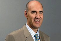 Headshot of a person smiling, wearing a beige suit, light blue shirt, and patterned tie against a plain background.