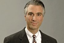 Headshot of a person in a suit and tie with gray hair, smiling slightly against a plain background.