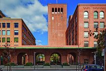 A historic brick building with arched windows and a central tower under a blue sky. The structure has a symmetrical design and an entrance with archways.