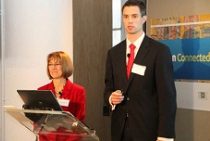 A small group of people in formal attire giving a presentation. One is standing behind a podium with a laptop, while another is holding a microphone.