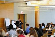 A person is speaking to a group in a conference or seminar setting. Attendees are seated at tables, and there are flip charts near the speaker.