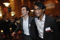 Two people in business attire are smiling and walking quickly in a lively and busy indoor setting, possibly an event or gathering.