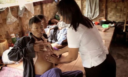 A healthcare worker is examining a person''s abdomen in a clinic or similar setting. Several individuals are resting in the background.