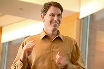 A person in a brown shirt smiling and gesturing energetically in a modern office setting.