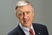 A headshot of a smiling, older adult wearing a suit with a red patterned tie against a plain background.