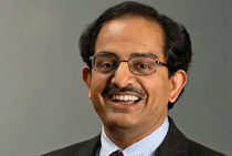 Headshot of a person with short dark hair, glasses, and a mustache, smiling and wearing a suit and tie against a gray background.