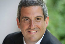 A headshot of a smiling person with short gray hair, wearing a dark suit, in front of a blurred natural background.