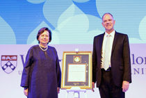 Two people standing beside a framed award or certificate on display, with university logos and a patterned background. The scene suggests a formal recognition or award ceremony.