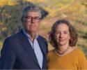 A couple posing outdoors with a blurred landscape background featuring autumn foliage. The individual on the left wears a suit and glasses, while the person on the right wears a yellow sweater and a necklace.