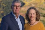 A couple posing outdoors with a blurred landscape background featuring autumn foliage. The individual on the left wears a suit and glasses, while the person on the right wears a yellow sweater and a necklace.