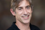 Headshot of a person smiling, with short grayish-brown hair, wearing a black shirt against a blurred background.