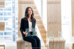 A person sitting indoors holding three glass awards, with additional awards placed beside them. The setting features a modern, artistic design with wooden slats and cityscape visible through large windows.