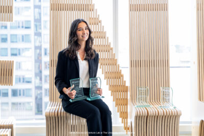 Young woman in business attire holding two glass awards, seated in a modern wodden bench with city views.