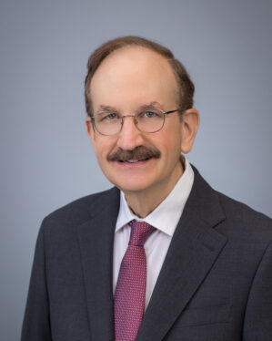 Headshot of a person with glasses and a mustache, wearing a dark suit, white shirt, and red tie, set against a plain background.